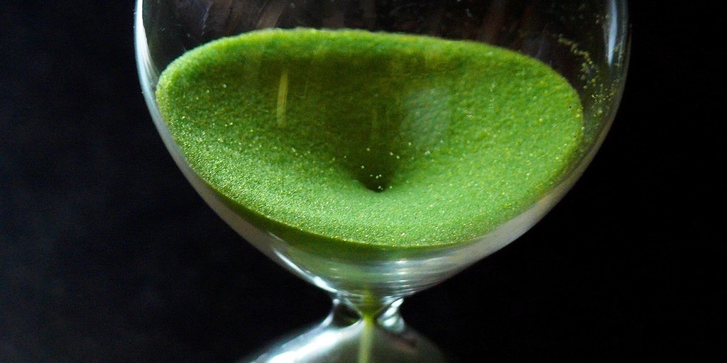 Close-up of an hourglass against black, its upper bulb full of bright green sand draining through the neck.